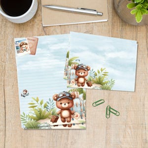 Op de afbeelding: Briefpapier set met een cartoonbeer met een pilotenmuts en koffer, tegen een tropische strandscène. De set bevat gelijnd papier, blanco kaarten en een paspoortillustratie. Groene paperclips en een koffiekopje zijn ook zichtbaar.