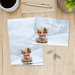 Op de afbeelding: Briefpapier-set met een illustratie van een Schotse hooglander met een winter sjaal en hulst, tegen een besneeuwd berglandschap. Het papier bevat horizontale blauwe lijnen om op te schrijven en wordt vergezeld door drie groene paperclips.