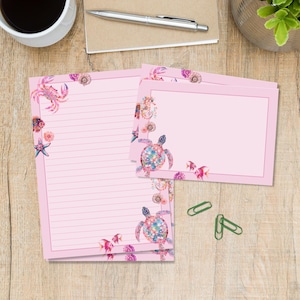 May include: A set of light pink stationery featuring lined and unlined paper decorated with colourful watercolour illustrations of sea turtles, crabs, starfish, fish, and seashells. Three green paper clips are placed next to the stationery.