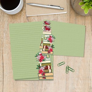 Op de afbeelding: Lichtgroen notitiepapier met horizontale lijnen en een boeken- en bloemenontwerp. Het ontwerp toont een stapel boeken versierd met rode poinsettia's en groene hulst. Twee groene paperclips zijn ook zichtbaar.