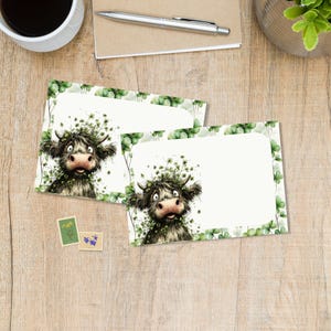 以下が含まれることがあります： 緑のクローバーのリースをつけたハイランド牛のイラストが描かれた、2枚の白紙のノートカード。カードは白い背景に緑のクローバーの縁取りがされています。切手も2枚見えます。