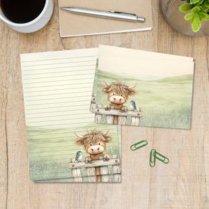 May include: Stationery set featuring a lined sheet and a matching envelope, both decorated with an illustration of a fluffy brown Highland cow leaning over a wooden fence in a green field with a small blue bird perched nearby.