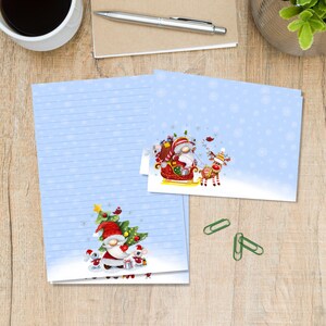 Op de afbeelding: Lichtblauwe briefpapier set met kerstthema illustraties. De set bevat gelijnd papier en kaarten met kabouters, rendieren en een kerstboom. Sneeuwvlokken zijn verspreid over de achtergrond, wat een feestelijke winterse scène creëert.
