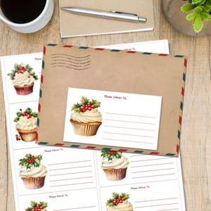 Peut inclure: Un ensemble de papeterie avec une enveloppe en papier kraft à rayures rouges et bleues, une étiquette d'adresse assortie et des autocollants de cupcakes. Le motif de cupcake comprend du glaçage blanc, du houx et des baies rouges. Un stylo et une tasse à café sont également visibles.