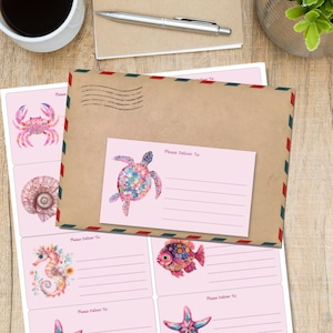 Può includere: Una busta di carta marrone con bordi da posta aerea rossi e verdi poggia su un foglio di etichette per spedizioni rosa chiaro. Le etichette presentano illustrazioni ad acquerello di una tartaruga marina, un granchio, una conchiglia, un cavalluccio marino.