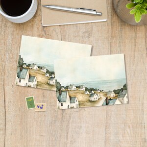 Peut inclure: Deux tirages d'art à l'aquarelle représentant un village côtier avec des maisons blanches, des toits gris et une mer bleue. Les tirages sont sur une surface en bois avec une tasse à café, un stylo, un bloc-notes et des timbres.