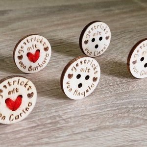 May include: Five wooden buttons with red yarn hearts and the text "gestrickt mit" and the name of the person who knitted the item. The buttons are on a light brown wooden surface.