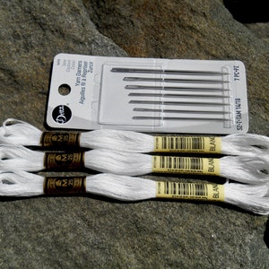 Op de afbeelding: Een verpakking met zeven garenstoppers en drie spoelen wit borduurgaren. De naalden zitten in een doorzichtige plastic verpakking. De spoelen garen hebben goudbruine etiketten met het woord "BLANC".