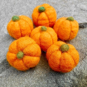 May include: Six felt pumpkins in orange with green stems. The pumpkins are arranged in a circle on a gray stone surface.