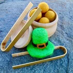 Può includere: Un cappello da folletto verde in feltro con una fibbia nera e arancione si trova su un vassoio di legno con due pinzette di legno. Un vaso di legno pieno di palline di feltro gialle si trova dietro il vassoio.