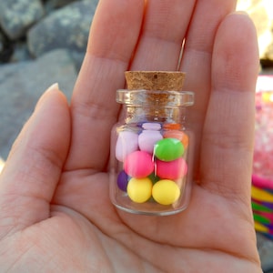May include: A small glass jar with a cork lid filled with colorful candy. The candy is in various shades of pink, purple, yellow, green, and orange.