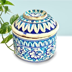 May include: A round, lidded ceramic container with a hand-painted floral design. The container features a cream-colored base with blue and white floral patterns and a matching lid. The design includes stylized flowers and leaf motifs, creating a decorative storage piece.