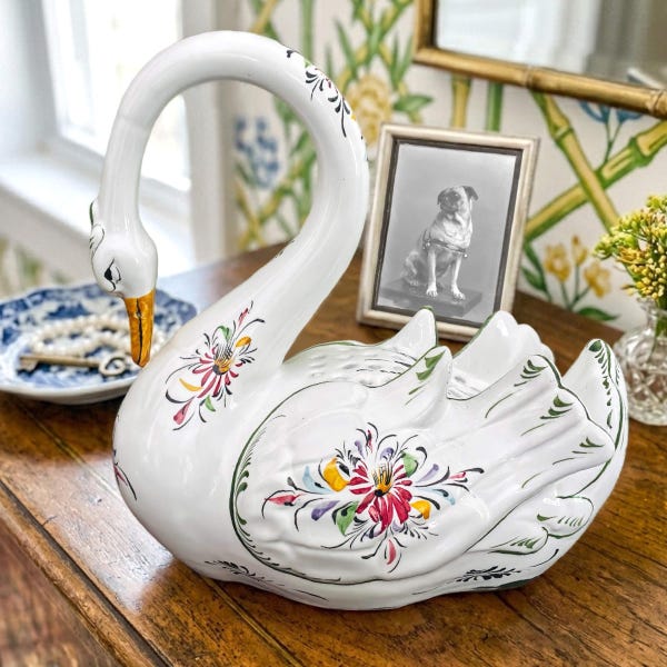 Może przedstawiać: Biała ceramiczna figurka łabędzia z ręcznie malowanymi motywami kwiatowymi w kolorze czerwonym, żółtym i zielonym oraz metalicznym złotym dziobem. Łabędź ma wygiętą szyję, szczegółowe pióra skrzydeł i wydrążony środek.