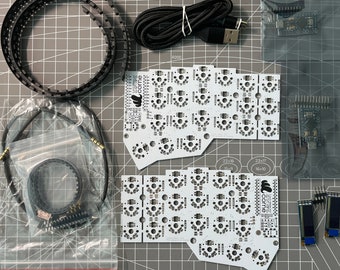 Puede incluir: Un conjunto de componentes electrónicos para construir un teclado. El conjunto incluye dos placas de circuito impreso, un cable, un cable USB, una bolsa de componentes electrónicos y dos pantallas pequeñas. Las placas de circuito impreso son blancas y tienen un diseño en blanco y negro. Las pantallas son negras y tienen una pantalla blanca.