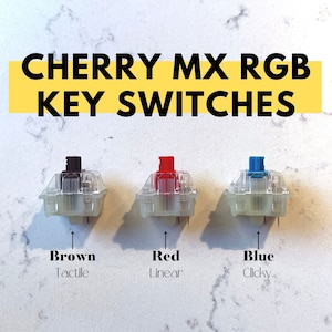 May include: Three Cherry MX RGB mechanical keyboard key switches. The switches are in clear housings with brown, red, and blue stems. The switches are labeled "Brown Tactile", "Red Linear", and "Blue Clicky".