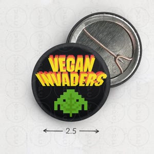 May include: A 2.5 inch button with a black background and the text "VEGAN INVADERS" in yellow, red, and orange. A green pixelated alien spaceship is below the text.