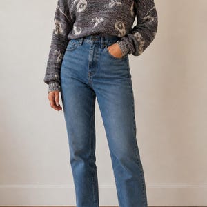 Könnte beinhalten: Ein langärmeliger Pullover mit Stehkragen in Dunkelanthrazit mit strukturiertem weißem abstraktem Muster, kombiniert mit einer mittelblauen Jeans im Straight-Leg-Schnitt mit klassischem Fünf-Taschen-Design, getragen mit beigen Wildleder-Slippern.
