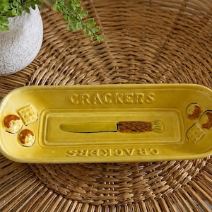 May include: A rectangular yellow ceramic serving tray with rounded corners. The word CRACKERS is embossed on the top and bottom edges. The center features a molded knife graphic, while the sides display raised images of crackers and cheese.