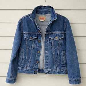 Puede incluir: Una chaqueta vaquera azul clásica con cierre de botones, dos bolsillos en el pecho y dos bolsillos laterales. La chaqueta está colgada de un gancho negro sobre un fondo de madera blanca. La chaqueta tiene cuello y puños abotonados.