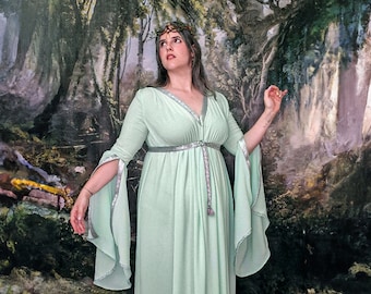 May include: A woman wearing a light green gown with silver trim and a silver belt. She is wearing a crown of leaves and flowers on her head. She is standing in front of a painting of a forest.
