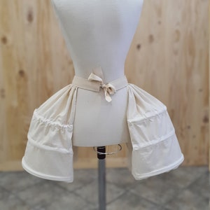 May include: A white cotton bustle with a gathered skirt and a tied sash. The bustle is on a white dress form.
