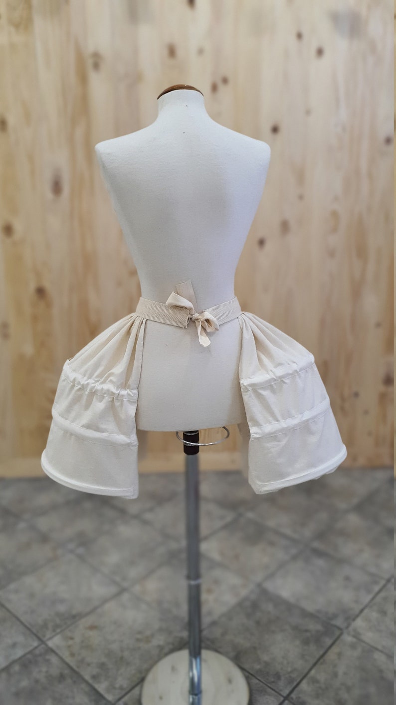 May include: A white cotton bustle with a gathered skirt and a tied sash. The bustle is on a white dress form.