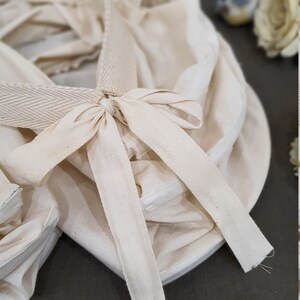 May include: A white cotton fabric basket with a white cotton ribbon tied in a bow. The basket is folded and has a textured surface.