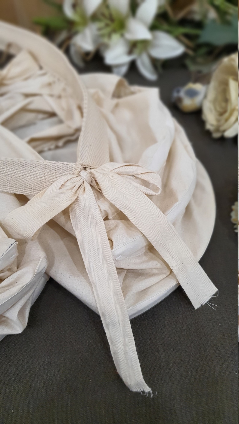 May include: A white cotton fabric basket with a white cotton ribbon tied in a bow. The basket is folded and has a textured surface.