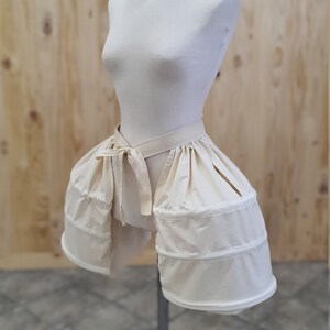 May include: A white cotton bustle with a gathered skirt and a tied sash. The bustle is on a white dress form.