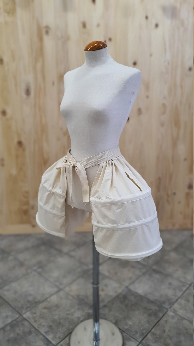 May include: A white cotton bustle with a gathered skirt and a tied sash. The bustle is on a white dress form.