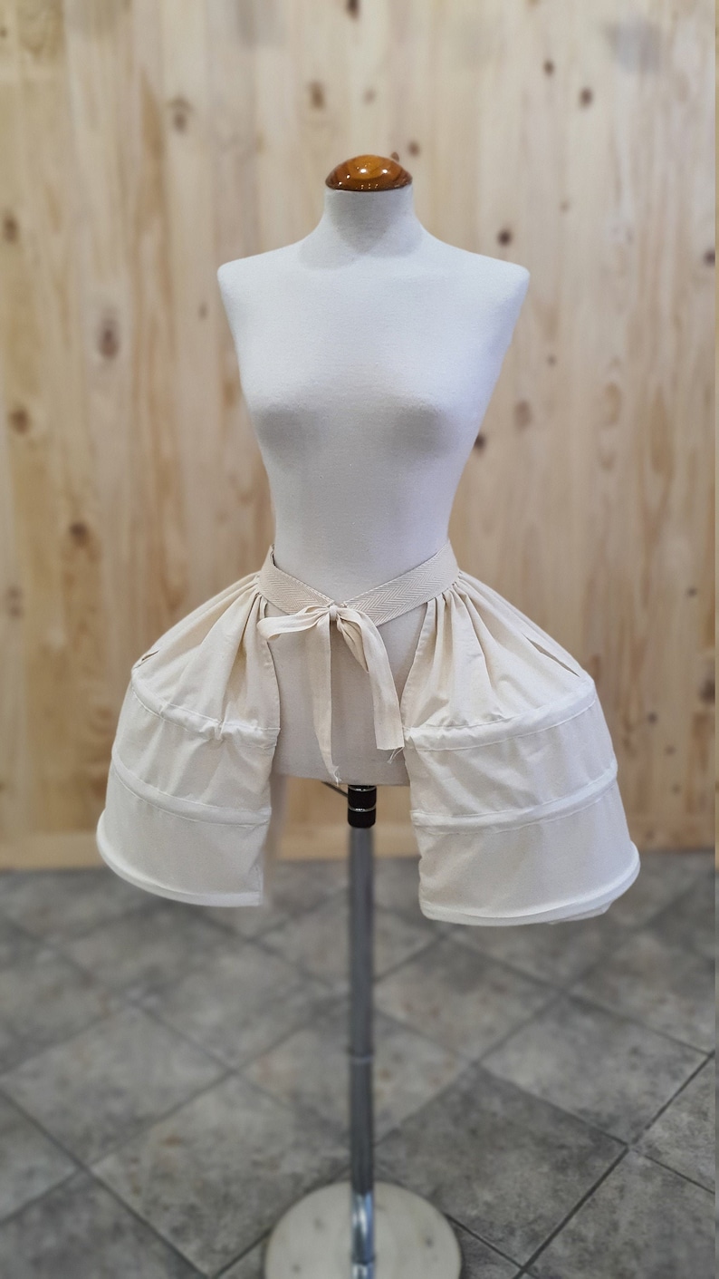 May include: A white cotton bustle with a tied ribbon at the waist. The bustle is on a white dress form.