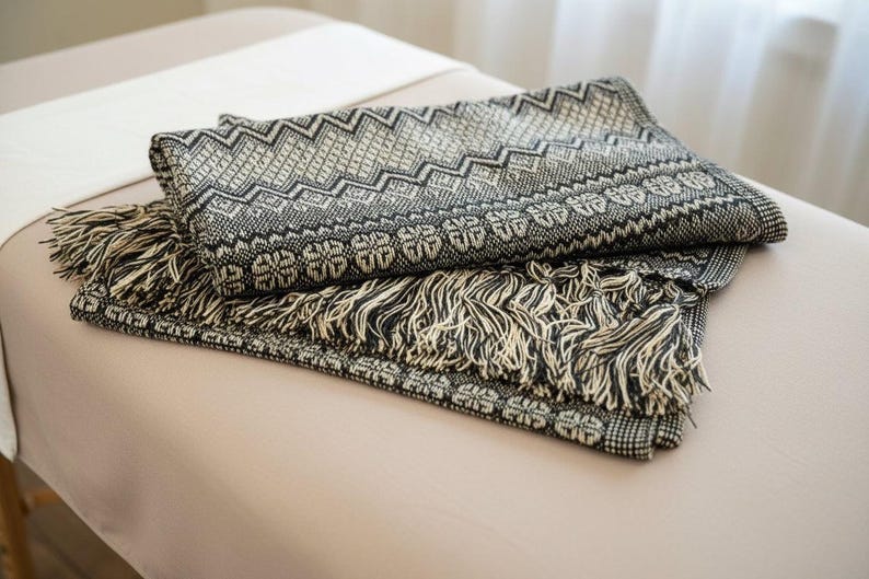 Puede incluir: Una manta plegada con estampado en blanco y negro con flecos. La manta tiene un dise&ntilde;o geom&eacute;trico con zigzags y patrones florales. La franja es de color crema. La manta est&aacute; sobre una superficie de color beige claro.