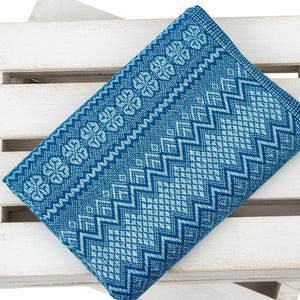 Puede incluir: Un textil azul doblado con un patr&oacute;n geom&eacute;trico de diamantes, zigzags y formas florales. El textil es de un tono azul medio con acentos azul claro. Est&aacute; sobre una caja de madera blanca.