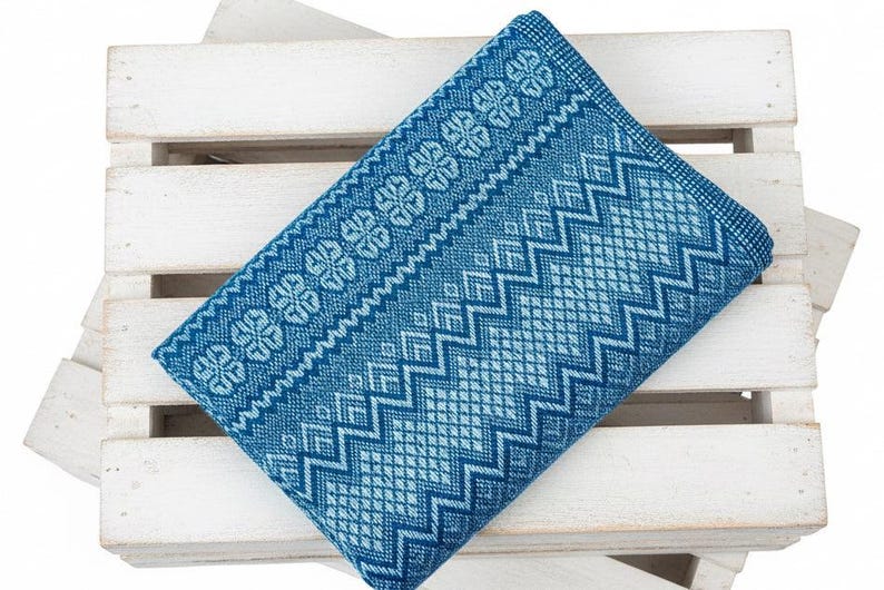 Puede incluir: Un textil azul doblado con un patr&oacute;n geom&eacute;trico de diamantes, zigzags y formas florales. El textil es de un tono azul medio con acentos azul claro. Est&aacute; sobre una caja de madera blanca.
