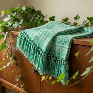 Puede incluir: Una manta tejida verde y blanca doblada con detalles de flecos. La manta est&aacute; sobre una c&oacute;moda de madera. Plantas de hiedra verde en el fondo y primer plano.