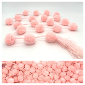 Könnte beinhalten: Eine Girlande aus hellrosa Pompons, verbunden durch einen passenden rosa Faden. Die Pompons sind flauschig und gleichmäßig verteilt, mit einer Quaste aus losen Fäden am Ende. Der Hintergrund ist weiß.