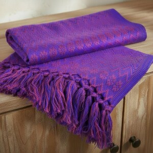 Puede incluir: Una manta tejida morada doblada con un estampado floral y flecos. La manta est&aacute; hecha de un material suave y se exhibe sobre un mueble de madera. Los flecos son una mezcla de morado y rosa.