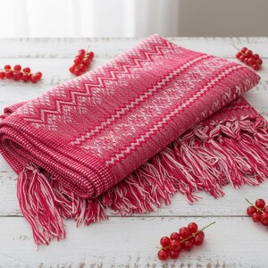 Puede incluir: Bufanda rosa y blanca plegada con un patr&oacute;n geom&eacute;trico y flecos. La bufanda est&aacute; rodeada de bayas rojas sobre una superficie de madera blanca. La bufanda tiene un patr&oacute;n en zigzag y diamante.