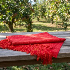 Puede incluir: Una bufanda roja doblada con bordes con flecos descansa sobre una mesa de madera desgastada. La bufanda tiene un patr&oacute;n tejido y es de un tono rojo vibrante. El fondo muestra un huerto borroso.