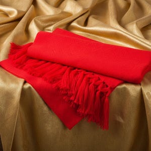 Puede incluir: Una bufanda roja vibrante con flecos est&aacute; colocada sobre una tela dorada y brillante. La bufanda est&aacute; doblada, mostrando su textura y dise&ntilde;o. El fondo es un material dorado rico, creando una sensaci&oacute;n de lujo.