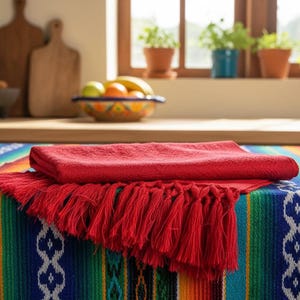 Puede incluir: Un vibrante y colorido camino de mesa serape con un mantel rojo con flecos. El camino de mesa presenta rayas azules, verdes, amarillas y naranjas. Un pa&ntilde;o rojo doblado descansa sobre el pa&ntilde;o con flecos, creando un efecto en capas.