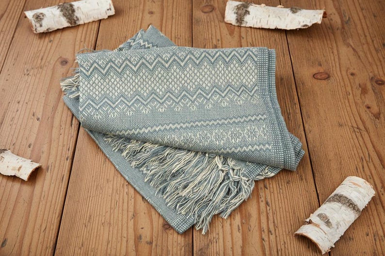 Puede incluir: Una manta plegada con estampado azul y blanco con flecos. La manta presenta un dise&ntilde;o geom&eacute;trico y est&aacute; hecha de un material suave. La manta se exhibe sobre una superficie de madera con detalles de corteza de abedul.