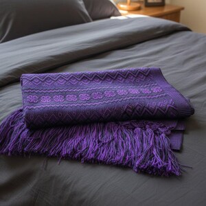 Puede incluir: Una manta morada doblada con un patr&oacute;n geom&eacute;trico y detalles de flecos descansa sobre una colcha gris. El dise&ntilde;o de la manta presenta motivos florales y en zigzag repetidos, agregando textura e inter&eacute;s visual a la ropa de cama.