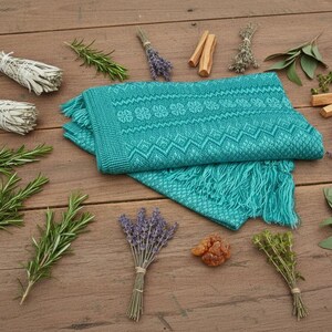 Puede incluir: Una bufanda tejida turquesa con un patr&oacute;n geom&eacute;trico y flecos est&aacute; rodeada de manojos de hierbas, salvia, romero y lavanda, junto con trozos de madera y resina, todo dispuesto sobre una superficie de madera.