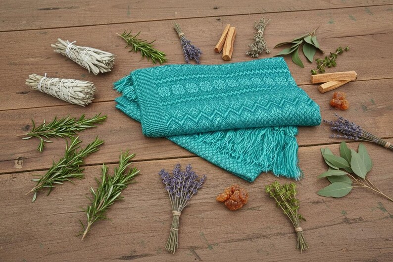 Puede incluir: Una bufanda tejida turquesa con un patr&oacute;n geom&eacute;trico y flecos est&aacute; rodeada de manojos de hierbas, salvia, romero y lavanda, junto con trozos de madera y resina, todo dispuesto sobre una superficie de madera.