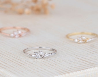 May include: Three delicate rings with leaf-like designs. The rings are in silver, rose gold, and gold. The silver ring in the foreground features a leaf design with small, sparkling stones. The rose gold and gold rings are slightly out of focus in the background.