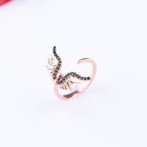 May include: A rose gold ring with black diamond wings. The wings are detailed with small black diamonds and the ring is open on one side.