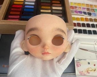 May include: A close-up of a doll head with painted-on eyebrows, eyelashes, and lips. The doll's eyes are open and have a realistic appearance. The doll's head is made of a light-colored material and is being held by a gloved hand.