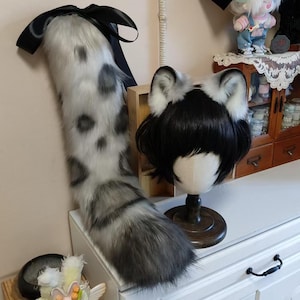 May include: A faux fur cat tail with a black ribbon and a matching set of cat ears on a wig stand. The tail is white with grey spots and stripes. The ears are white with grey tips and black hair.