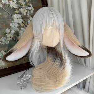 以下が含まれることがあります： 長い白髪と大きなベージュと白のウサギの耳が付いたコスプレウィッグ。ウィッグには、お揃いのベージュと白の尻尾も付いています。耳の内側はピンク色で、先端は茶色です。ウィッグはマネキンの頭に表示されています。